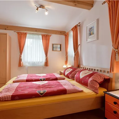 Ein gemütliches Schlafzimmer mit zwei Betten, roten Bettwäsche und sanften Vorhängen. An der Wand hängen Bilder, und es gibt einen kleinen Nachttisch mit einer Lampe.