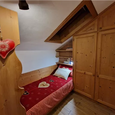 A cozy bedroom with wooden furniture and a red bed. A heart-shaped decoration hangs on the wall.