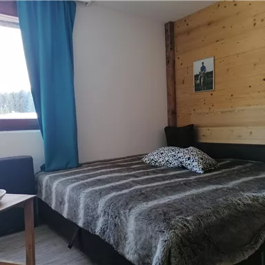 A cozy room with a double bed and a wooden wall. Next to the bed is a small table with a bowl of oranges and a window with a view.