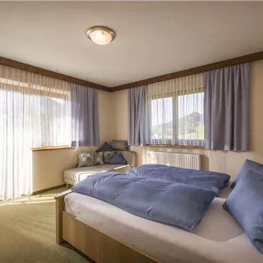 Ein gemütliches Schlafzimmer mit einem großen Bett und blauen Kissen. Helle Vorhänge lassen Tageslicht herein und bieten einen Blick auf die umliegende Landschaft.