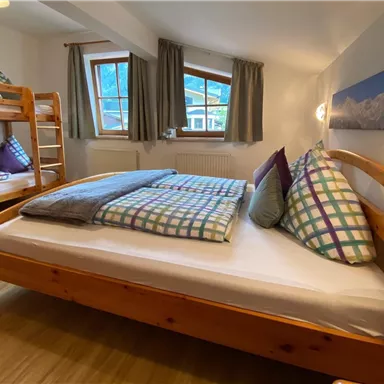 Ein gemütliches Zimmer mit einem Doppelbett und einem Etagenbett. Die Wände sind hell, und große Fenster sorgen für viel Tageslicht.