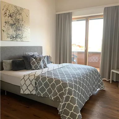 Ein modernes Schlafzimmer mit einem großen Bett und dekorativen Kissen. Große Fenster lassen viel Tageslicht hinein und zeigen einen ansprechenden Holzboden.