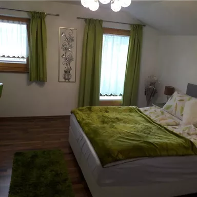 A cozy bedroom with a large bed and green bedding. Green curtains and a small desk create an inviting atmosphere.