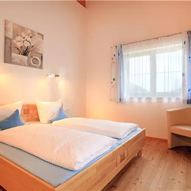 A cozy bedroom with a double bed and soft lighting. The room has a window with curtains and a comfortable chair in the corner.
