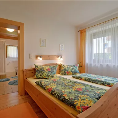 A cozy bedroom with a double bed and colorful bedding. The large windows let in plenty of light and offer a view outside.