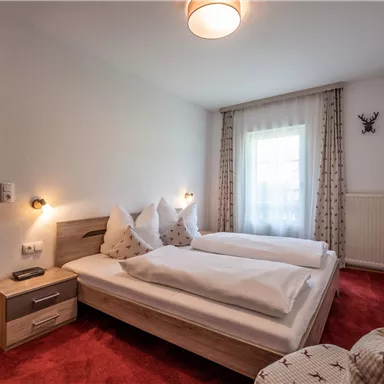 A cozy bedroom with two single beds and red carpets. Large windows provide plenty of daylight and an inviting atmosphere.