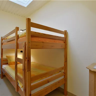 Ein einfaches Zimmer mit einem Hochbett aus Holz und zwei Matratzen. Ein Stuhl und eine kleine Pflanze sind ebenfalls vorhanden.