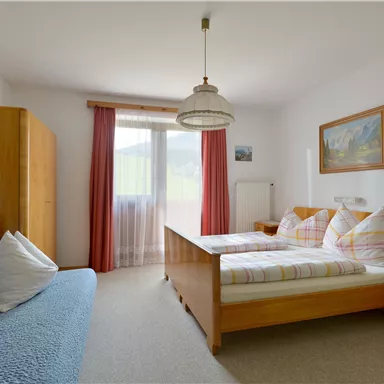 Ein helles Schlafzimmer mit zwei Betten und einem Sofa. Große Fenster mit natürlichem Licht und eine warme, einladende Einrichtung.