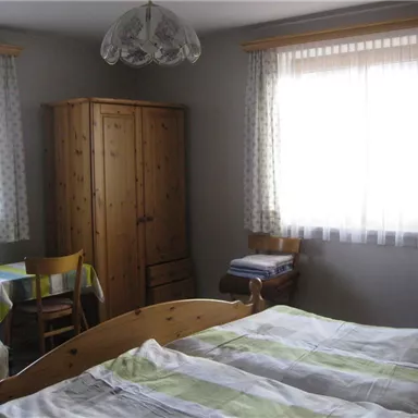 A simple bedroom with two single beds and a wooden wardrobe. The room has large windows with curtains and a small table with a chair.