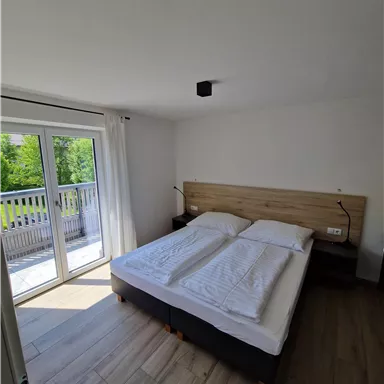 Ein helles Schlafzimmer mit einem Doppelbett und frischen Bettwäsche. Große Fenster bieten einen Blick auf die Natur draußen.