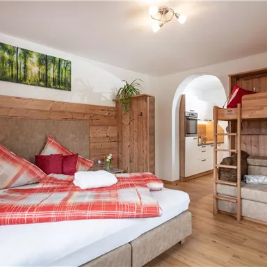 A cozy bedroom with a large bed and colorful pillows. In the background, a small kitchen and a loft bed are visible.