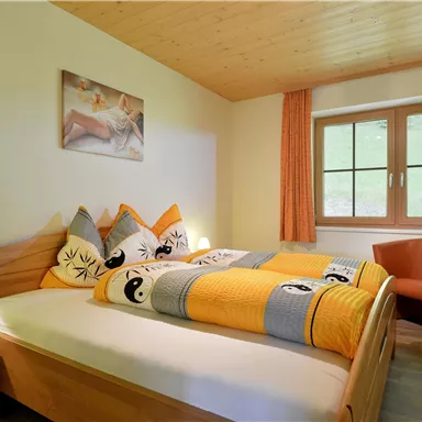 A cozy bedroom with a double bed and colorful pillows. The room has large windows and warm wooden decor.