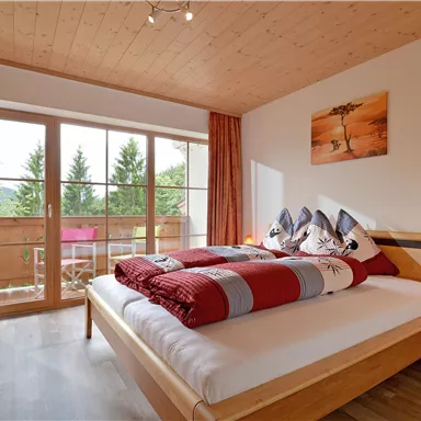 A cozy bedroom with a large bed and colorful blankets. The room has large windows overlooking a balcony and green trees.