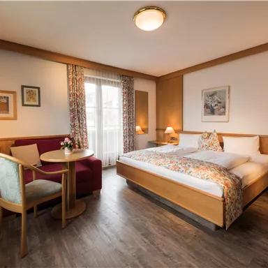 A bright and cozy hotel room with a large bed and wooden paneling. A table with chairs and windows with curtains complete the furnishings.