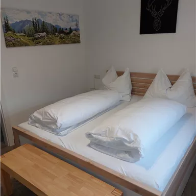 A cozy bedroom with a double bed, two pillows, and a wooden bench. A landscape painting and an artwork featuring a deer motif hang on the wall.