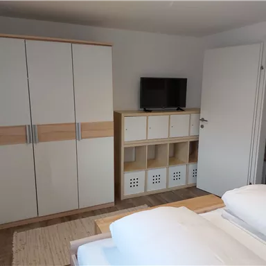 A simple bedroom with two beds, a wardrobe, and a television. On the wall, there is a shelf with storage space.