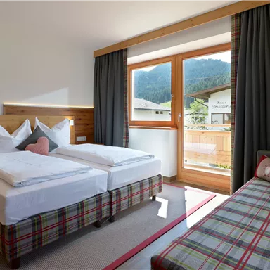 A bright hotel room with two beds and a cozy sofa. Large windows provide a view of the mountains and let in plenty of daylight.