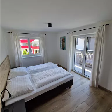 Ein helles Schlafzimmer mit einem Doppelbett und modernen Möbeln. Große Fenster bieten viel Tageslicht und Zugang zum Balkon.