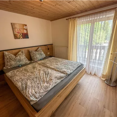 Ein gemütliches Schlafzimmer mit einem großen Doppelbett und schönen Kissen. Durch die Fenster strömt Tageslicht und es gibt einen kleinen Balkon.
