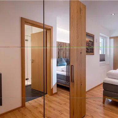 A modern bedroom with wooden furniture and a large mirror. In the background, a comfortable bed and a door to a bathroom are visible.