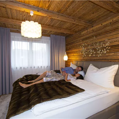 Ein gemütliches Schlafzimmer mit Holzverkleidung und modernem Design. Ein Paar liegt entspannt im großen Bett unter einer warmen Decke.
