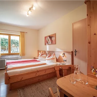 Ein helles Zimmer mit zwei Betten und einem kleinen Tisch. Vor dem Fenster sind Vorhänge und eine Blumendekoration auf dem Tisch.