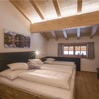 Ein gemütliches Schlafzimmer mit Holzdecke und drei Betten. Große Fenster bieten viel Tageslicht und einen Blick nach draußen.