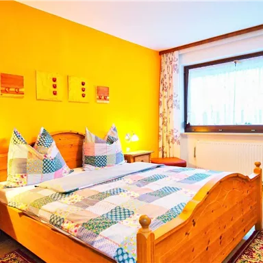 Ein gemütliches Schlafzimmer mit einem großen Doppelbett und bunten Kissen. Die Wände sind in warmem Gelb gestrichen und es gibt einen roten Sessel neben einem Fenster.