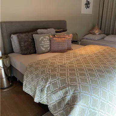 Ein gemütliches Schlafzimmer mit einem Bett, das mit einer dekorativen Decke und mehreren Kissen geschmückt ist. An der Wand hängen ein paar dekorative Elemente, während ein leichter Teppich den Raum ergänzt.