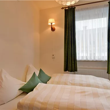 The image shows a cozy bedroom with two beds and green cushions. Bright curtains hang in front of the window, letting in daylight.