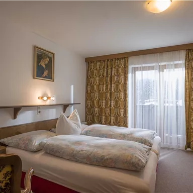 Ein gemütliches Schlafzimmer mit zwei Betten und einer warmen Farbgestaltung. Große Fenster sorgen für Tageslicht und Blick nach draußen.