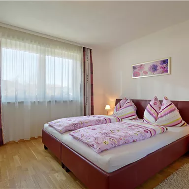 A cozy bedroom with two beds and colorful bedding. Large windows provide plenty of daylight.