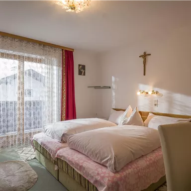 Ein gemütliches Schlafzimmer mit zwei Betten und sanften Farben. Große Fenster mit Vorhängen sorgen für viel Tageslicht.