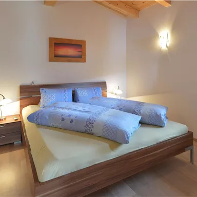 A cozy bedroom with a large bed and wooden ceiling. The room is bright and friendly, with soft colors and a window.