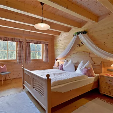 Ein gemütliches Schlafzimmer aus Holz mit einem Doppelbett und rosa Kissen. Große Fenster lassen viel Licht herein und schaffen eine einladende Atmosphäre.