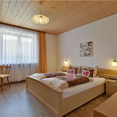 A cozy bedroom with a double bed and heart-shaped decorative pillows. The room has large windows, bright wood, and a friendly atmosphere.