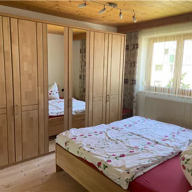 Ein helles Schlafzimmer mit einem großen Bett und einem Kleiderschrank aus Holz. Durch das Fenster fällt Tageslicht in den Raum.
