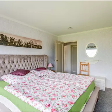 Ein gemütliches Schlafzimmer mit einem großen, gepolsterten Bett und bunten Bettbezügen. Die Wände sind hell und es gibt ein runde Spiegel sowie ein Bild von Bergen.