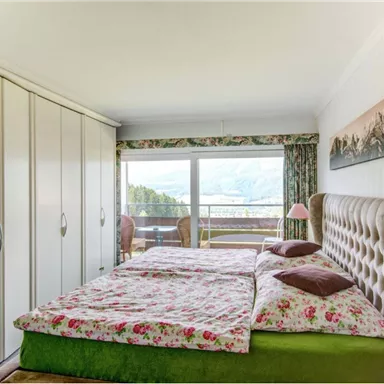 Ein gemütliches Schlafzimmer mit einem Doppelbett und bunten Bettwäsche. Große Fenster bieten einen Blick auf die Natur und viel Tageslicht.
