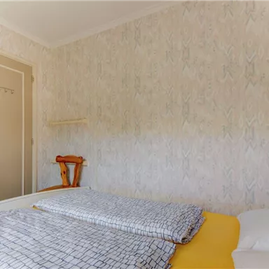Ein einfaches Schlafzimmer mit einem Bett und einer gelben Bettdecke. Die Wände sind mit einem hellen, gemusterten Papier dekoriert, und eine Tür führt nach draußen.