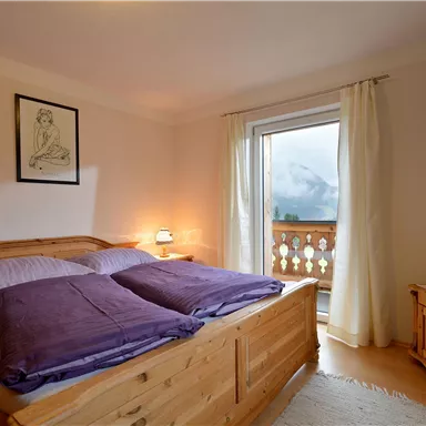 A cozy bedroom with a wooden bed and purple pillows. Large windows offer a view of the mountains.