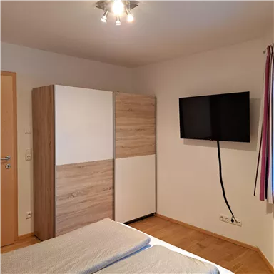 A modern bedroom with a double bed, a wardrobe, and a TV on the wall. The room has a bright and cozy atmosphere.