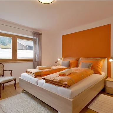 Ein gemütliches Schlafzimmer mit einem großen Bett und orangefarbenen Akzenten. Große Fenster sorgen für viel Tageslicht und eine angenehme Atmosphäre.