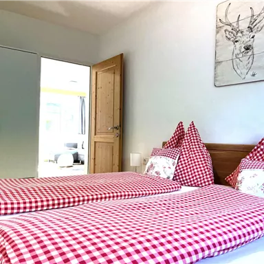 A cozy bedroom with two single beds, outfitted with checkered bedding in red and white. A picture of a deer hangs on the wall, and the room feels bright and inviting.