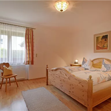 A cozy bedroom with a double bed and wooden furniture. The room has large windows, curtains in warm colors, and a carpet on the floor.
