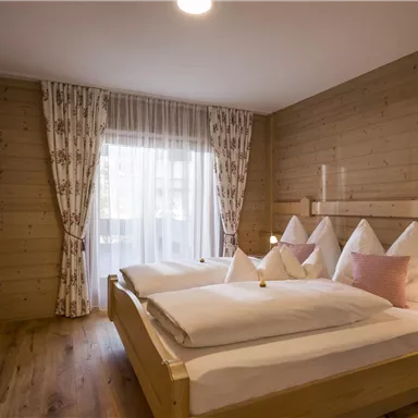 A cozy bedroom with two single beds and wooden paneling. Large windows with curtains let in plenty of light.