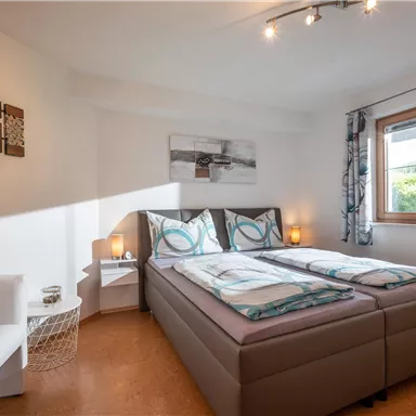 A modern bedroom with a double bed and a cozy armchair. The room has large windows and a bright, inviting atmosphere.