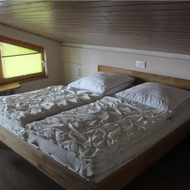 A cozy bedroom with a double bed and two large pillows. The windows are equipped with green blinds, providing daylight.