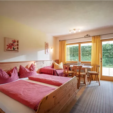 Ein gemütliches Zimmer mit einem Doppelbett und pinken Kissen. Es gibt einen Esstisch und große Fenster, die auf einen gepflegten Garten blicken.