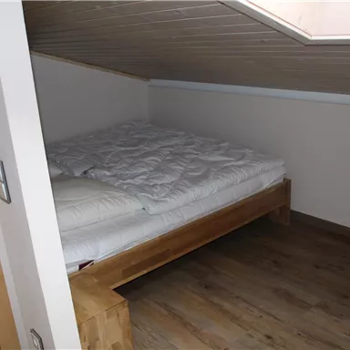 A cozy bedroom with a wooden bed and a mattress. The room has a slanted ceiling and a warm wooden floor.